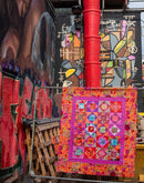 A design from the book Kaffe Fassett's Quilts in London showing colorful quilt hanging against a vibrant mural with red pipes.