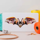 Mandala Bat Cross Stitch design upright on table top surrounded by thread, scissors, and hoop.