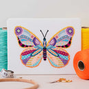 Mandala Butterfly Cross Stitch design upright on table surrounded by thread, scissors, hoop, and measuring tape.