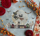 Night Riders cross-stitched pattern of a witch flying on a broomstick with a moon and village scene, surrounded by autumn leaves and flowers.