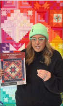 Person holding a quilt pattern book in front of a colorful quilt