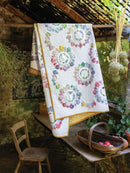 Alice's Country Diary Quilt showing colorful floral wreath motifs on a white background hanging from a rustic wooden beam in an outdoor garden setting with a table, chair, and hanging herbs.