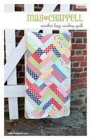 Another Lazy Sunday quilt with geometric pattern on a white stand against a brick wall.