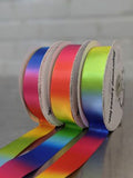 Three rolls of rainbow-colored ribbon arranged in a row, with the ribbons unspooled in front. The spools show a variety of color transitions including pink, yellow, green, blue, and red, set against a neutral gray background.  Double-Face Satin Ribbon 22mm (.875") Wide in Rainbow