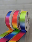 Three rolls of rainbow-colored ribbon arranged in a row, with the ribbons unspooled in front. The spools show a variety of color transitions including pink, yellow, green, blue, and red, set against a neutral gray background.  Double-Face Satin Ribbon 22mm (.875") Wide in Rainbow