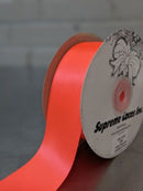 A close-up of a spool of bright orange satin ribbon with a white label reading 'Supreme Laces Inc.' on a dark surface. Double-Face Satin Ribbon 38mm (1.5") Wide