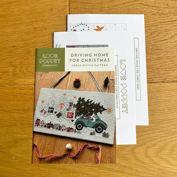 Cross stitch pattern called Driving Home for Christmas has a Christmas theme on a wooden surface, featuring 'Love Poppet' branding.