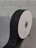 A roll of black glitter ribbon with reflective speckles, shown partially unrolled from a white spool on a gray surface against a light gray brick wall background.  Glitter Polypropylene Ribbon 38mm (1.5") Wide