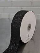 A roll of black glitter ribbon with reflective speckles, shown partially unrolled from a white spool on a gray surface against a light gray brick wall background.  Glitter Polypropylene Ribbon 38mm (1.5") Wide