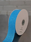 A spool of bright blue glitter ribbon on a dark gray surface with a light gray brick wall background. Glitter Polypropylene Ribbon 38mm (1.5") Wide
