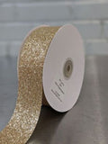 A roll of glittery gold ribbon on a white spool, photographed on a dark tabletop with a light gray brick wall in the background. Glitter Polypropylene Ribbon 38mm (1.5") Wide