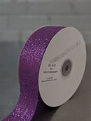A spool of glittery purple ribbon with a white cardboard core, shown on a gray surface against a light gray brick wall background. Glitter Polypropylene Ribbon 38mm (1.5") Wide