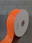 A roll of bright orange glitter ribbon with a white cardboard core, shown with a strip unspooled on a dark surface against a light gray brick wall background.  Glitter Polypropylene Ribbon 38mm (1.5") Wide