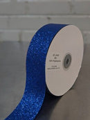 A roll of glittery royal blue ribbon on a white spool, resting on a gray surface with a white brick wall in the background.  Glitter Polypropylene Ribbon 38mm (1.5") Wide