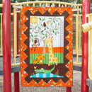A colorful quilt called Go Bananas eaturing safari animals and a tree, hanging outdoors on a playground structure with red and yellow metal bars.