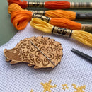 Wooden Hedgehog Needle Minder e cross-stitch fabric and colored threads on a light surface.