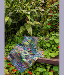 A colorful patchwork quilt with intricate patterns in shades of blue, green, and purple, draped over a dark wooden bench in a lush garden setting.  From the book, "Kaffe Fassett's Quilts at an English Manor House".