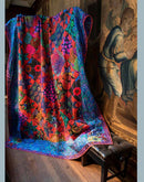 A large, vibrant quilt with a colorful floral and geometric pattern draped over a black leather stool in a traditional room with wooden floors and a classic painting on the wall.  Frpm the book, "Kaffe Fassett's Quilts at an English Manor House".