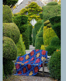 A colorful  quilt with a blue base and red, yellow, and orange star patterns draped over a bench in a garden topiary pathway from the book, Kaffe Fassett's Quilts at an English Manor House.