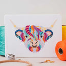 Mandala Highland Cow Cross Stitch design upright on table top surrounded by thread, scissors, and embroidery hoop.