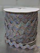 A spool of wavy, zigzag rick rack trim with a multicolor iridescent glitter finish, shown on a neutral gray background. Metallic Iridescent Rick Rack 15mm (.625") Wide