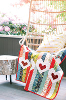 Pattern from "Not Your Typical Jelly Roll Quilts" shows colorful quilt with heart patterns draped over a chair on a patio with flowers in the background.