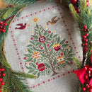 Embroidered Oh Christmas Tree Cross Stitch Pattern with festive elements on fabric, surrounded by greenery and berries.