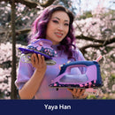 Yaya Han holding colorful irons with cherry blossom trees in the background, featuring the name Yaya Han.