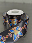 A roll of black satin ribbon with a colorful butterfly pattern in blue, purple, and gold, displayed on a neutral gray surface. Printed Single-Face Satin Ribbon 38mm (1.5") Wide - Butterfly on Black