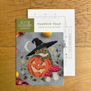 Cross stitch pattern for 'Pumpkin Toad' with a frog in a witch hat and pumpkin design, on a wooden surface.