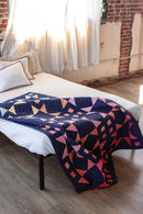 Pattern in "Quilts in Modern Color" shows geometric patterned quilt on a bed with a brick wall background.