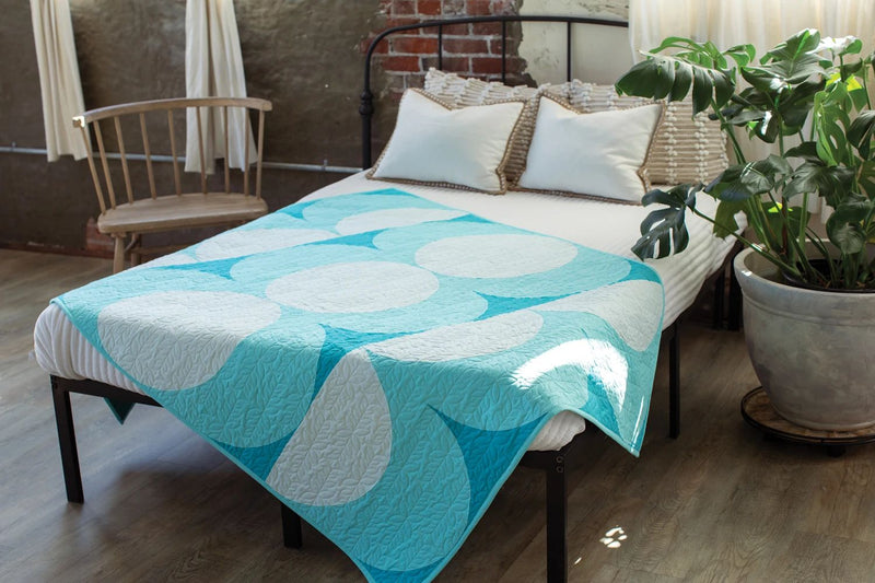 Pattern in "Quilts in Modern Color" shows  bed with a blue and white patterned quilt in a room with a plant .and chair.