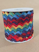 A spool of rick rack ribbon in a rainbow of colors, including red, orange, yellow, green, blue, and purple, wound on a white cardboard core against a neutral tan background. Rainbow Rick Rack 15mm (.625") Wide