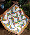 A colorful quilted blanket or throw called Rainbow Swirls, draped outdoors on a forest floor.