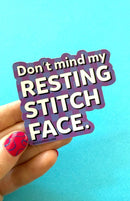 Sticker with text 'Don't mind my resting stitch face' held by a hand with pink nail polish on a blue background.