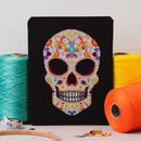 Skull Candy Cross Stitch design upright on tabletop, surrounded by thread, scissors, and hoop.