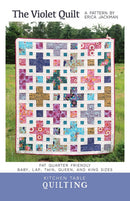 Front cover of 'The Violet Quilt' quilting pattern  by Erica Jackman, featuring a colorful patchwork quilt displayed outdoors on grass with trees in the background, and a blue band at the bottom reading 'Kitchen Table Quilting'.